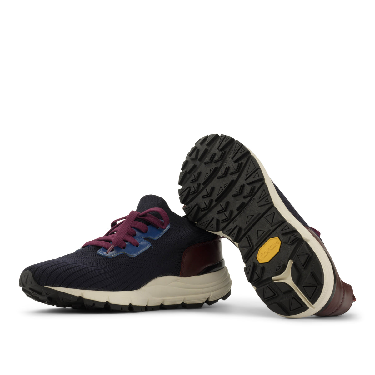 A pair of athletic trail running shoes shown on a white background, with one shoe standing upright to display the side profile and the other lying on its side to show the treaded outsole. The shoes feature a dark navy blue knit upper, burgundy laces, a blue accent near the laces, a cream midsole, and a small yellow Vibram logo on the outsole.