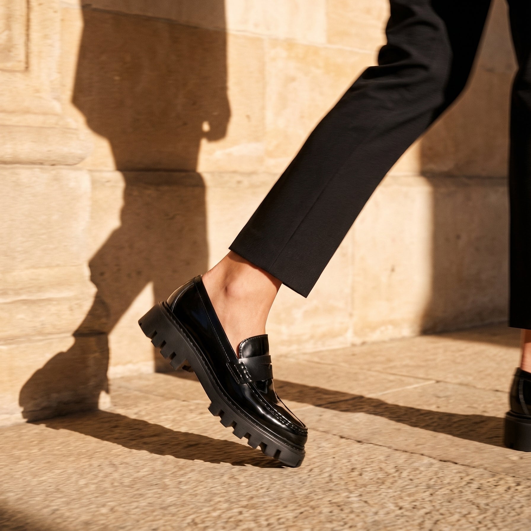 A person wearing black leather penny loafers with chunky lug soles and black trousers, walking outdoors against a sunlit beige stone wall.